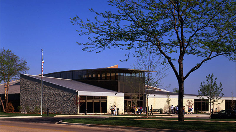 Modern building with a sloped roof, glass windows, and an American flag outside, surrounded by trees and a clear sky.