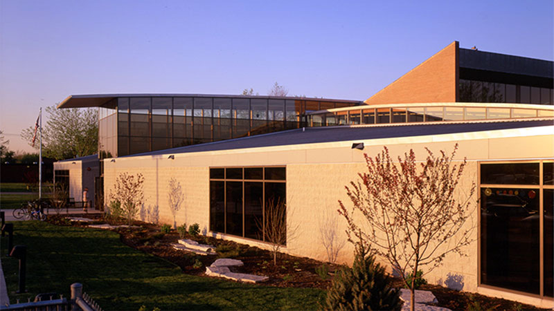 Modern building with glass windows and geometric rooflines, surrounded by landscaped greenery and a blue sky.