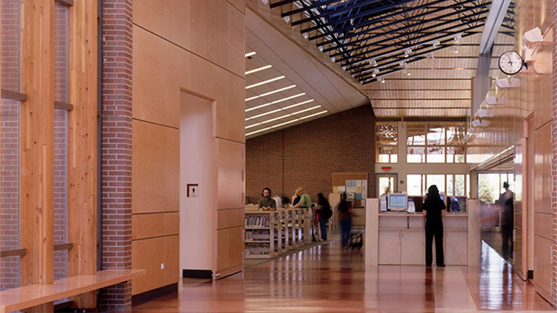 Wood-paneled library interior with computers, bookshelves, and people near the entrance under a high, angled ceiling.