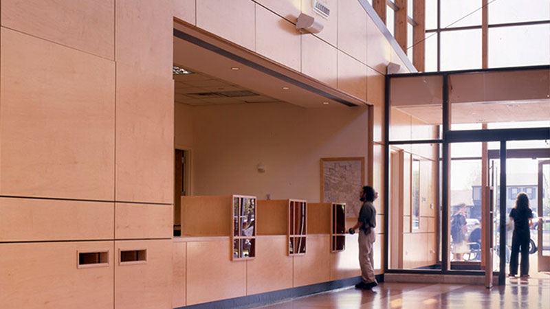 Reception area with light wood paneling, wall-mounted literature racks, and glass entrance. Two people are present.