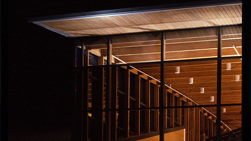 Exterior view of a modern building at night with large glass panels and wooden ceiling under a flat, overhanging roof.