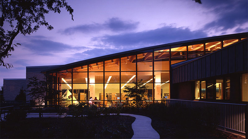 Illuminated modern building with curved glass windows at dusk, featuring a path leading towards the entrance.
