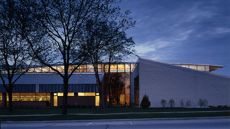 A modern library building with large windows, illuminated at dusk, surrounded by trees and situated near a quiet street.