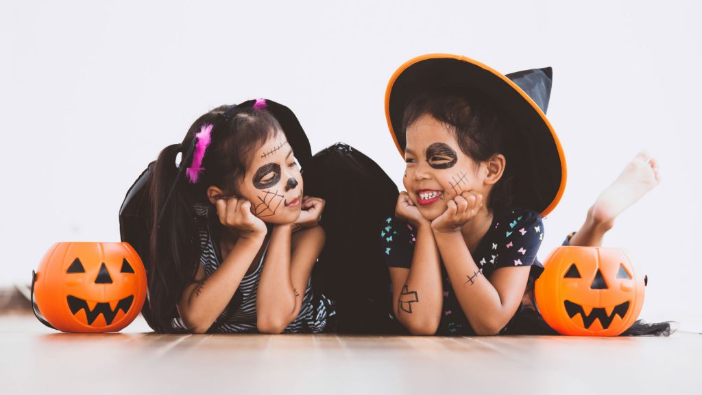 Two children in Halloween costumes and face paint lying on the floor, smiling with jack-o'-lantern buckets beside them.