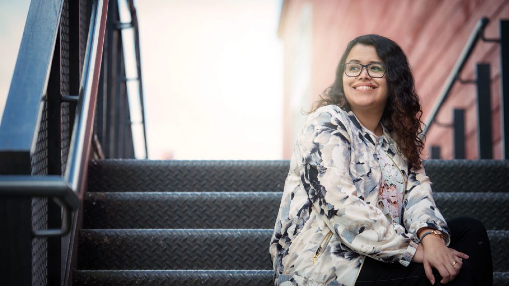 A smiling woman with glasses and a floral jacket sits on outdoor stairs, looking off into the distance.