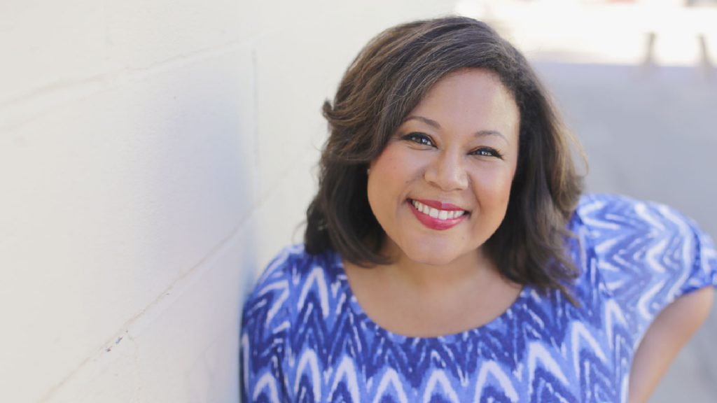 A person with shoulder-length hair smiles, wearing a blue and white zigzag patterned top, leaning against a wall.