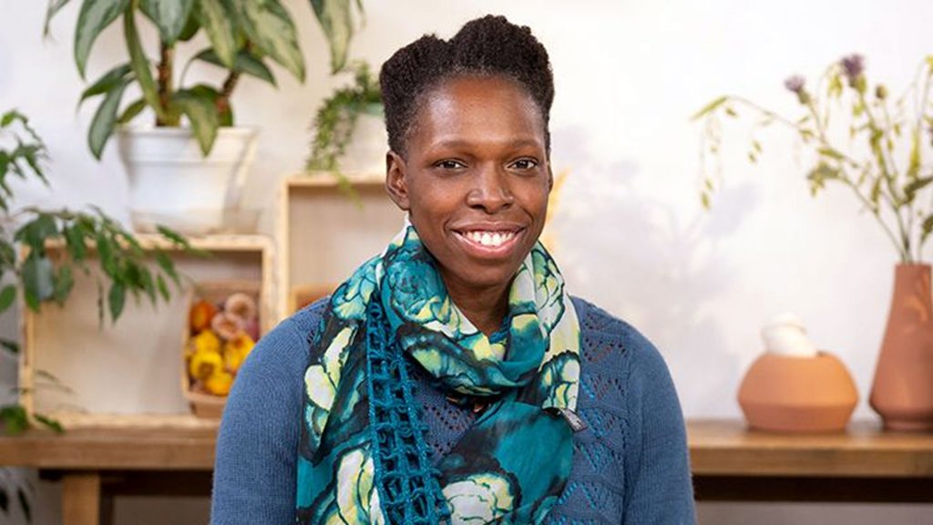 Smiling person in a blue sweater and patterned scarf, with plants in the background, posing indoors.