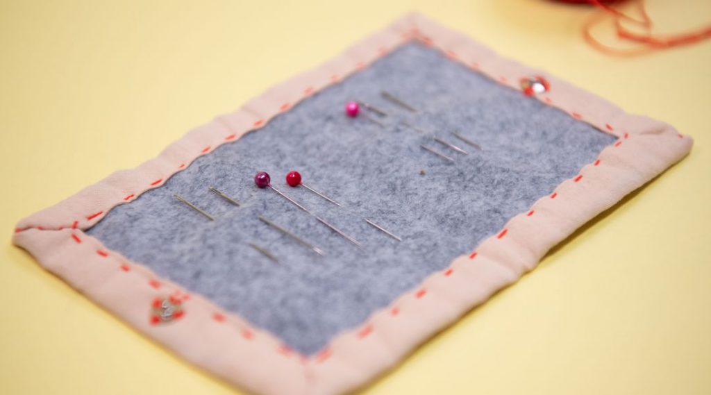A felt needle holder with multiple sewing needles, including two with pink heads, placed on a light yellow surface.