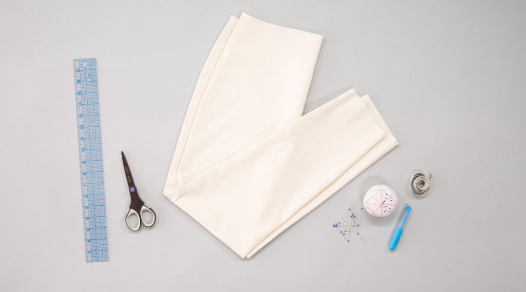 Sewing materials on a gray surface: fabric, scissors, ruler, pins, pin cushion, seam ripper, and measuring tape.