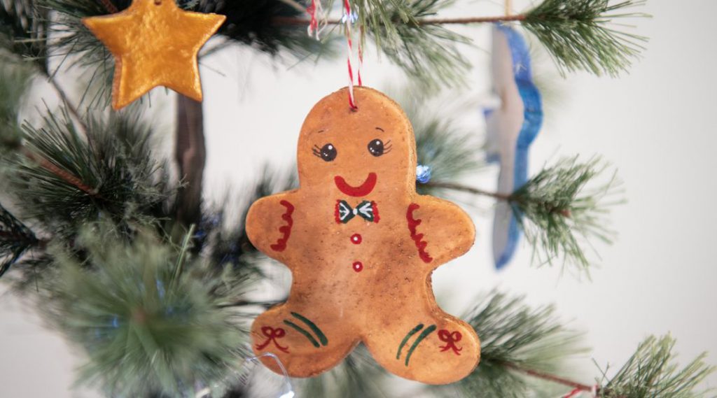 A gingerbread man ornament with a bow tie is hanging on a Christmas tree decorated with other ornaments.