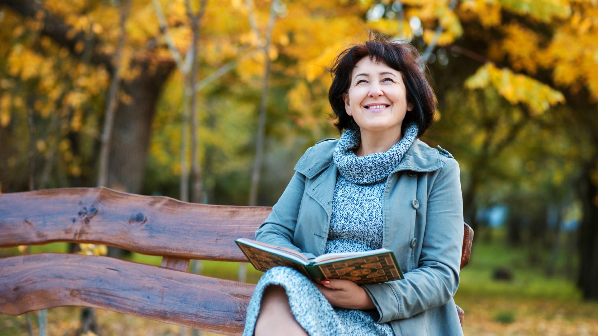 A woman sits on a park bench with a book, smiling and enjoying the autumn scenery with colorful leaves in the background.