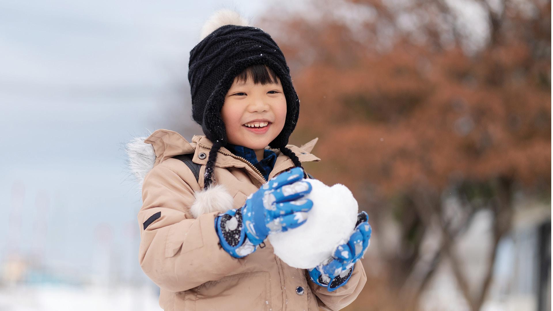 Child in winter clothes joyfully holds snowball outdoors.