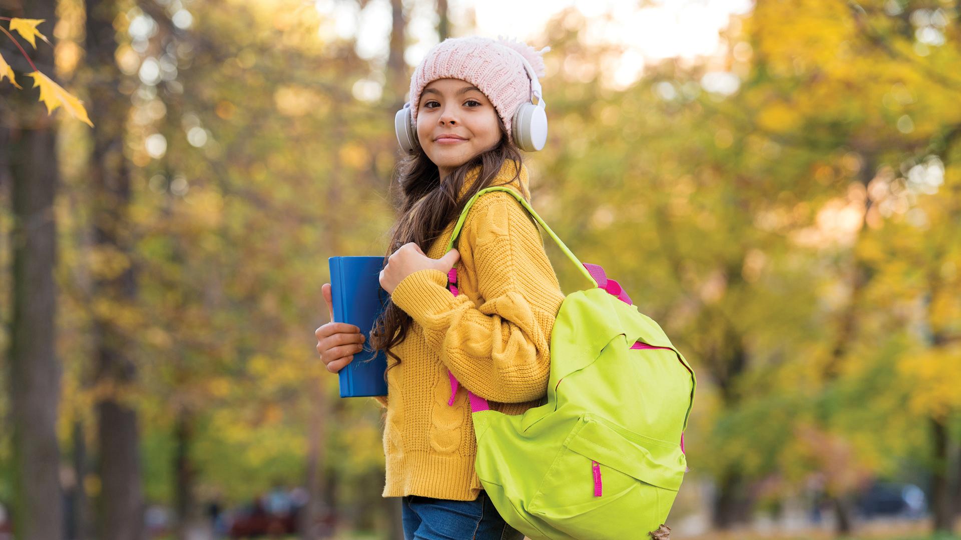 Smiling girl with headphones, yellow sweater, and backpack holds a book in a park with autumn trees.