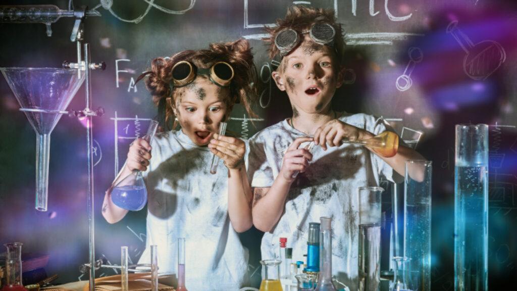 Two kids in goggles with messy hair, looking surprised while experimenting with colorful liquids in a chemistry lab.