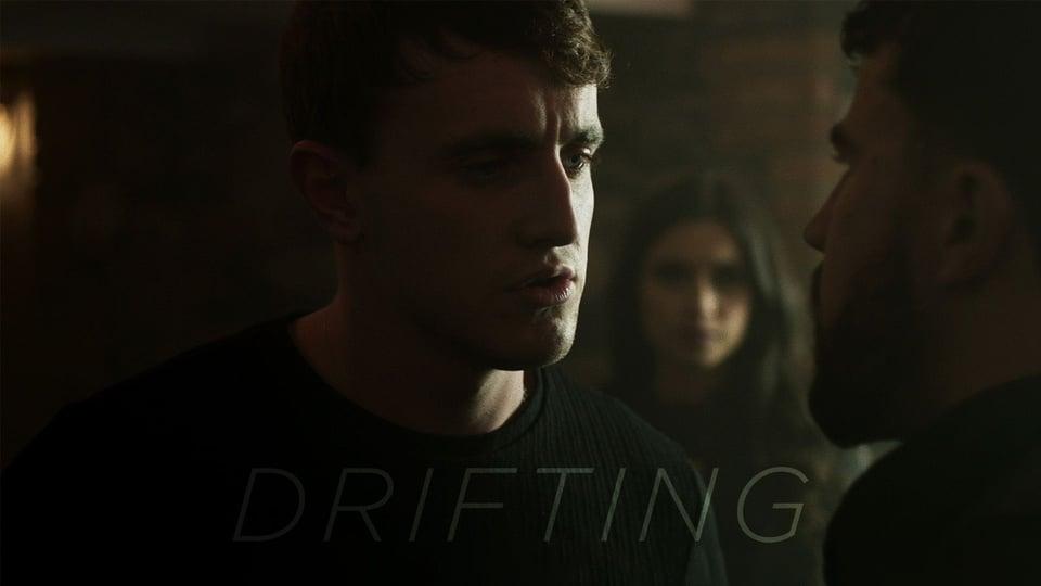 A tense scene with three people; a man in the foreground faces another, while a woman watches in the background. "Drifting" text.