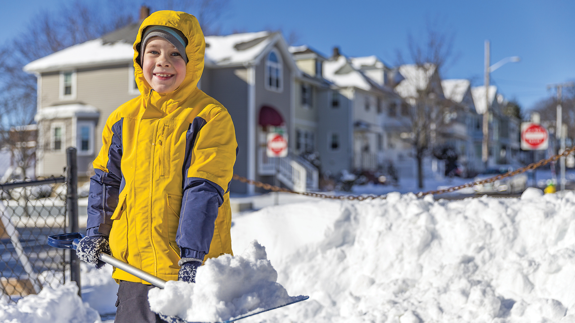 Smiling child in a yellow jacket shovels snow outside on a sunny winter day in a neighborhood.