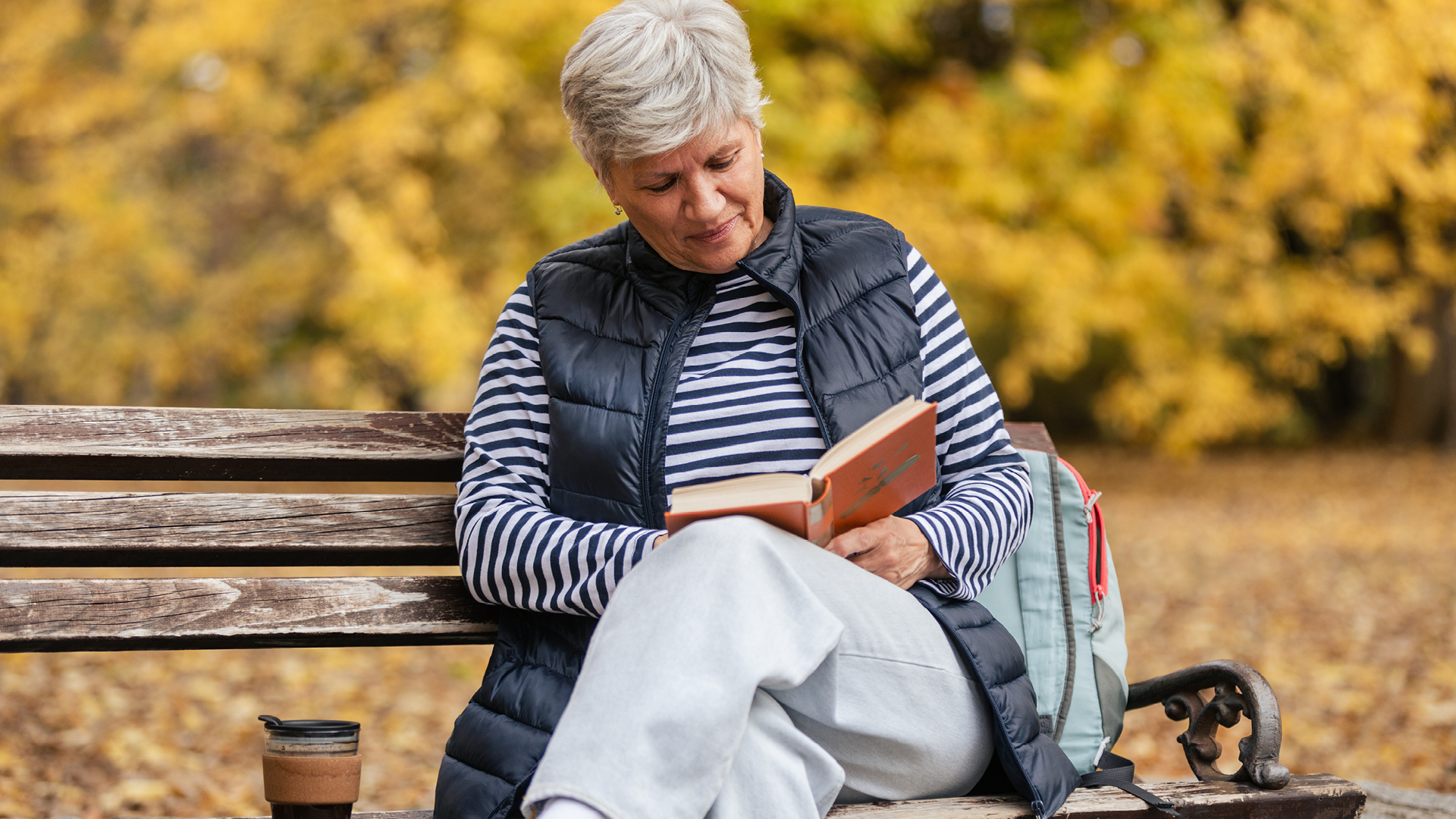 Older woman reading a book on a park bench in autumn, with coffee and a backpack beside her.