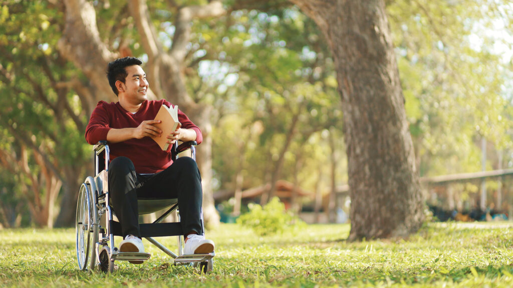 Man in wheelchair holding a book, smiling and looking away while sitting outdoors in a sunny park.