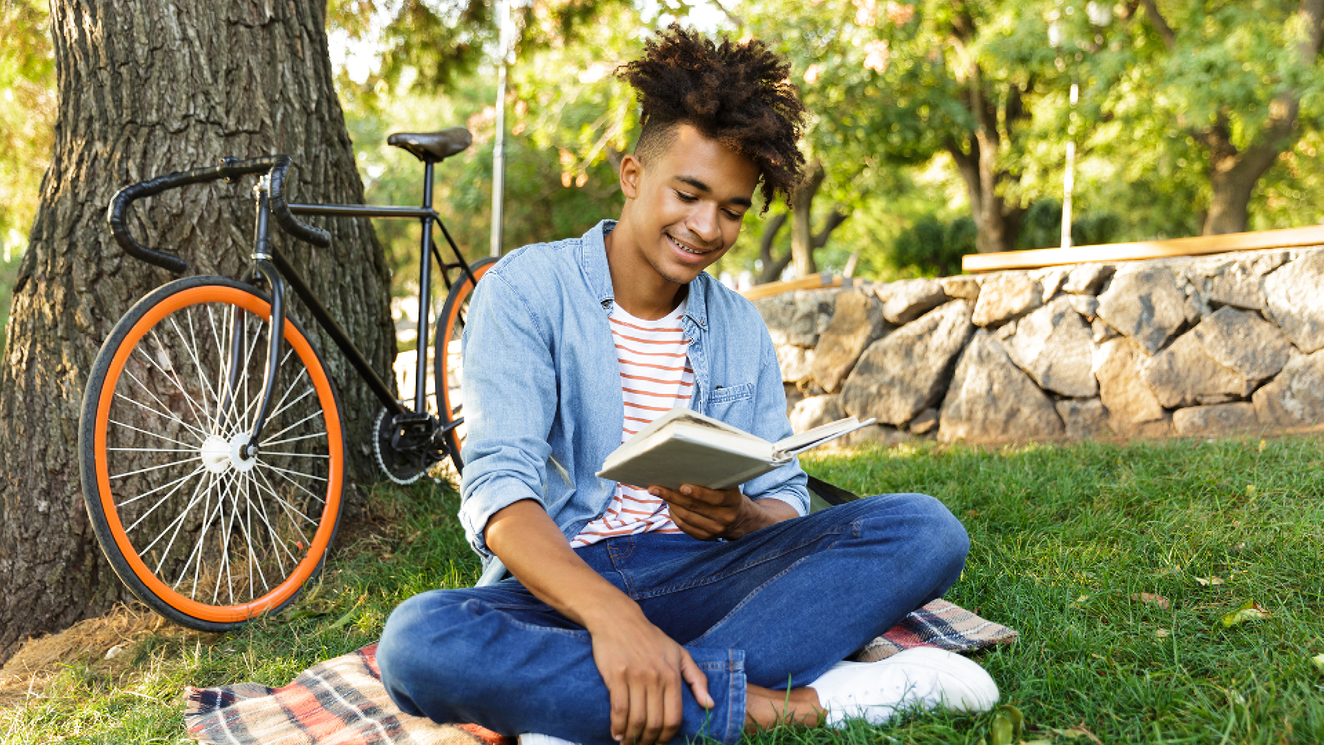 Smiling person sits on a blanket in a park, reading a book with a bicycle parked nearby.