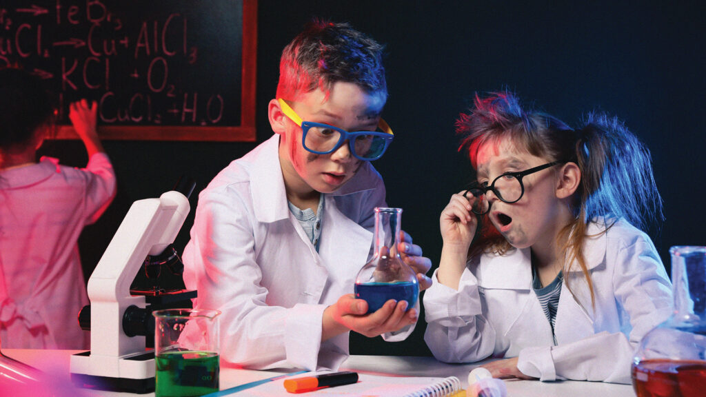 Two children in lab coats and goggles experiment with a beaker of blue liquid in a science classroom.