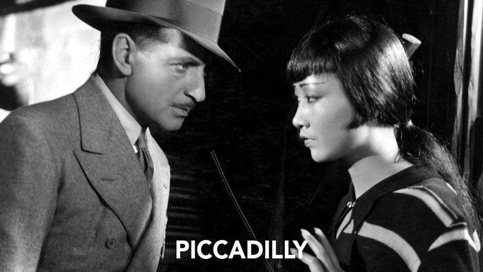 A man in a suit and hat speaks to a woman with bangs; text reads “PICCADDILY.” Black-and-white film scene.