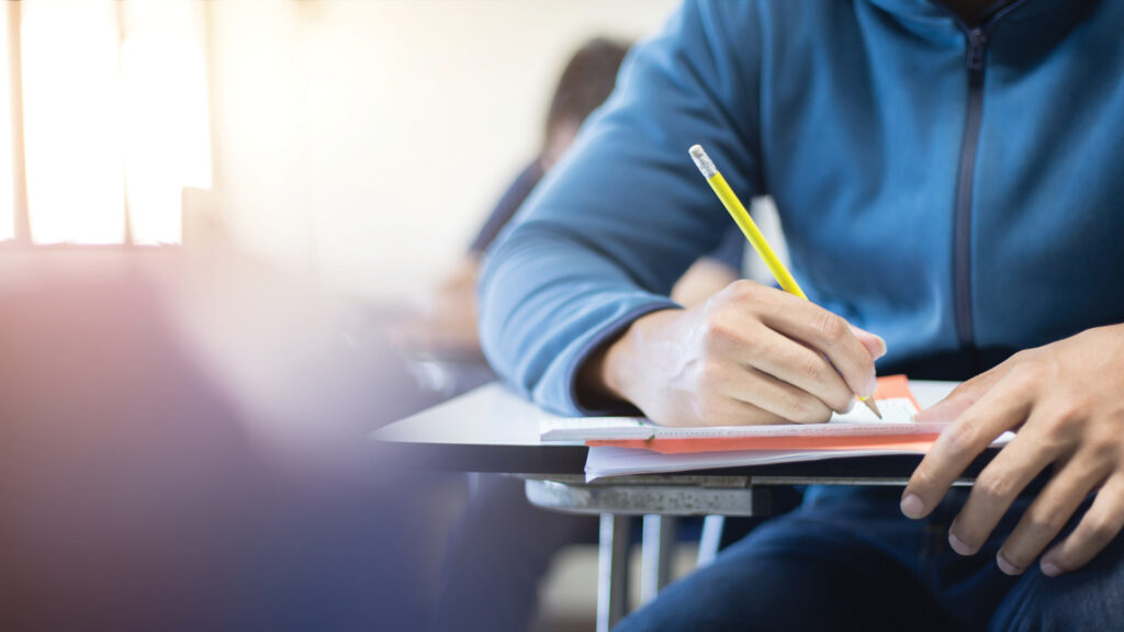 Student in blue hoodie writing with pencil on paper at a desk in a classroom setting.
