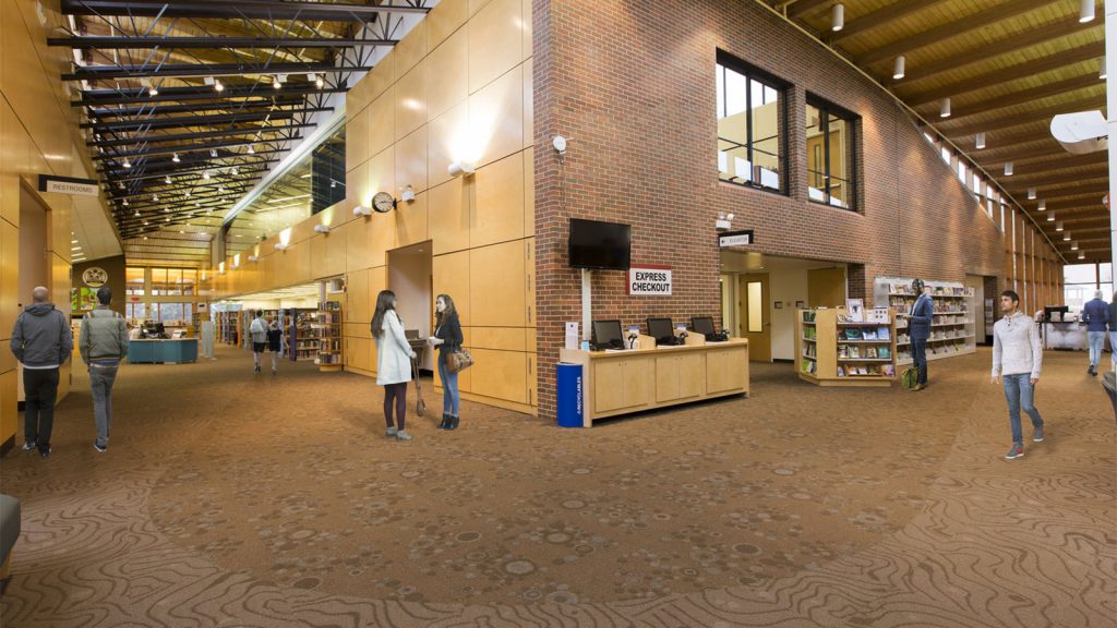 Library interior with people standing and walking, bookshelves, a checkout counter, and large windows in the background.