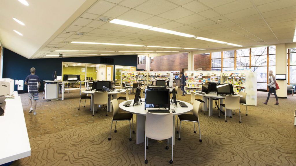 A spacious library with computer stations, book shelves, and people walking and browsing.