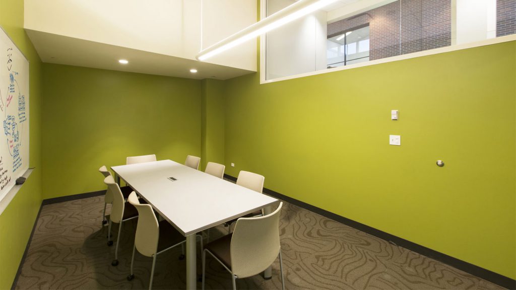 A small conference room with a white table, six chairs, a whiteboard on the wall, and green painted walls.