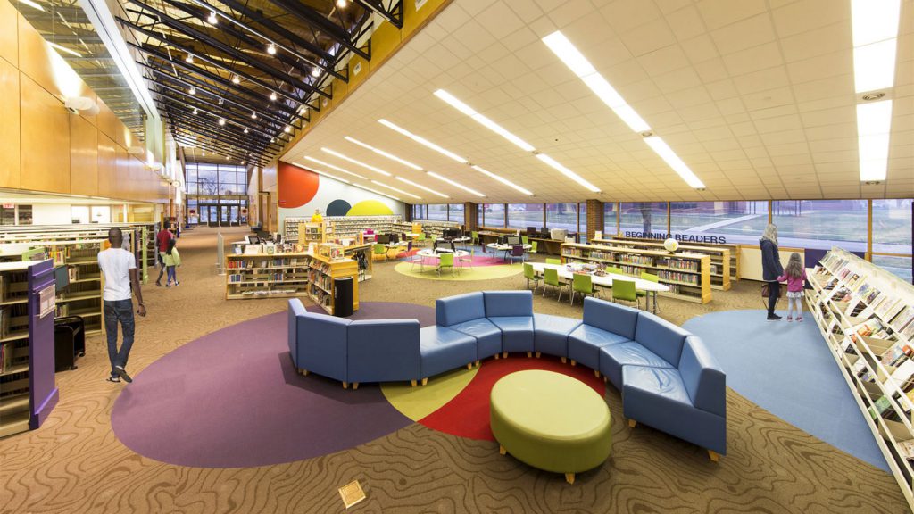 A spacious, modern library with colorful furniture, bookshelves, and a reading area under a bright ceiling.