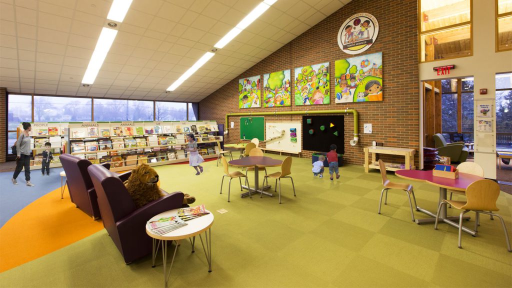 Children's section of a library with colorful murals, bookshelves, seating, activity tables, and a play area on the wall.