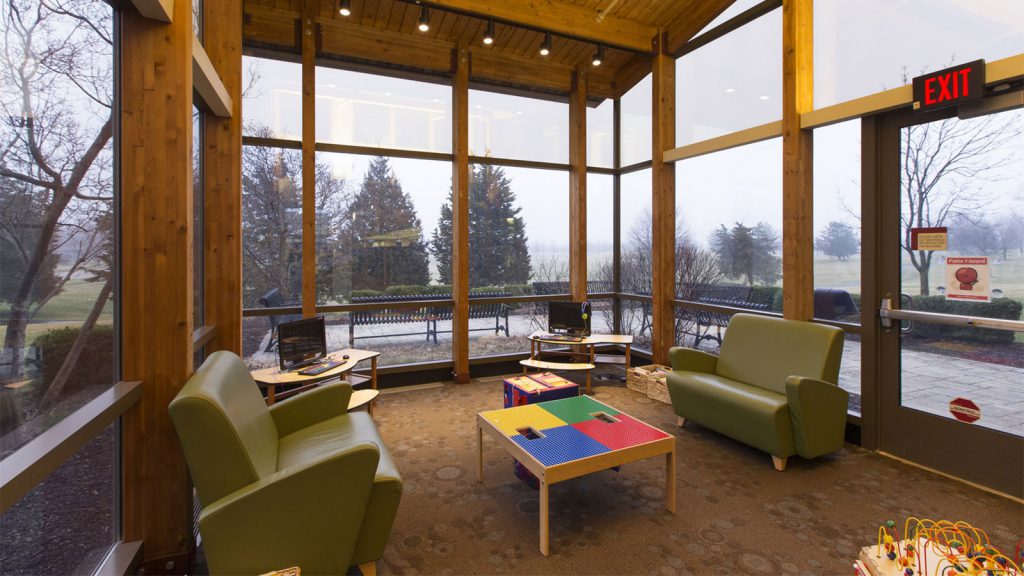 Children's playroom with green couches, low tables, toys, and a large window with a view of trees and overcast skies.