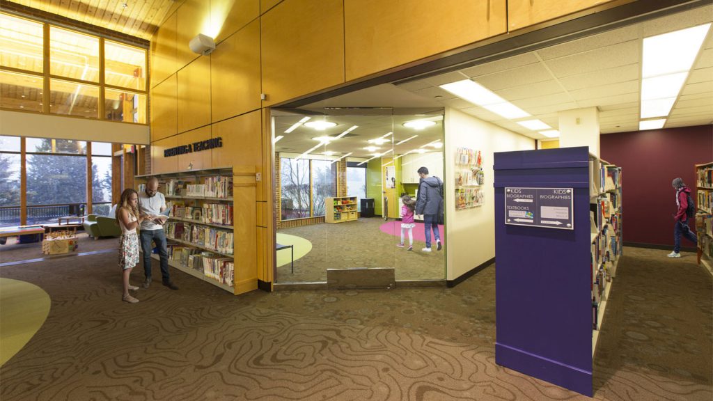 People explore a brightly lit library with wood and glass elements, looking at books and interacting with displays.