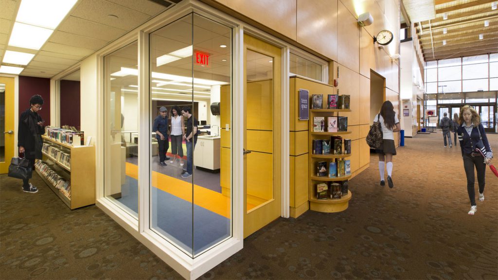 People walk in a library near a glass room with bookshelves and a clock on the wall.