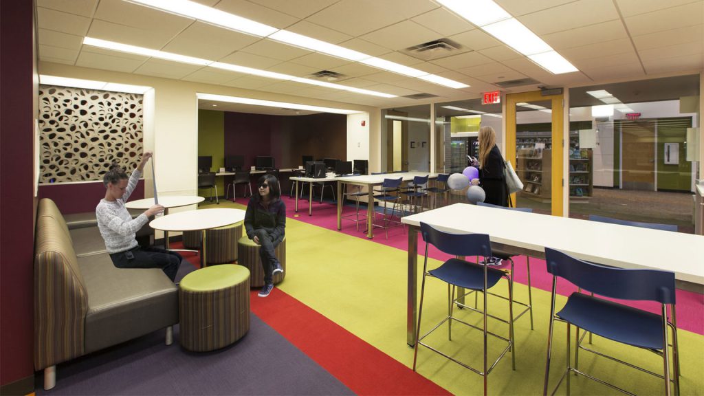 A modern, colorful office lounge area with people sitting and standing, featuring tables, chairs, and a computer station.