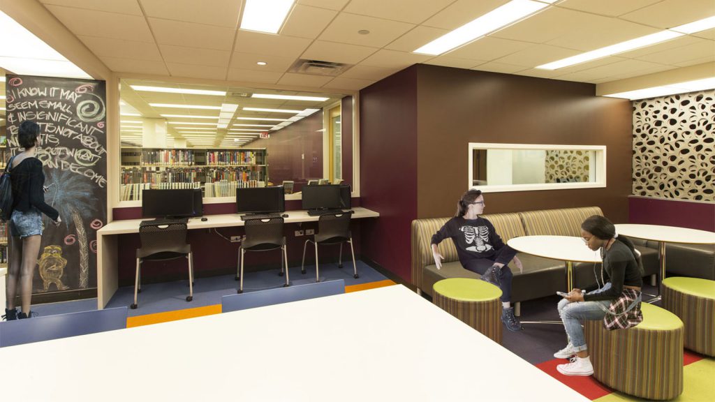 Modern library space with computers, a chalkboard wall, shelves of books, and two people sitting and talking at a table.