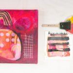 Abstract painting with bold pinks, reds, and geometric patterns next to a paintbrush and palette.