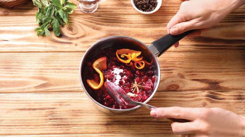 Hands stirring cranberries, orange slices, and spices in a saucepan on a wooden table.