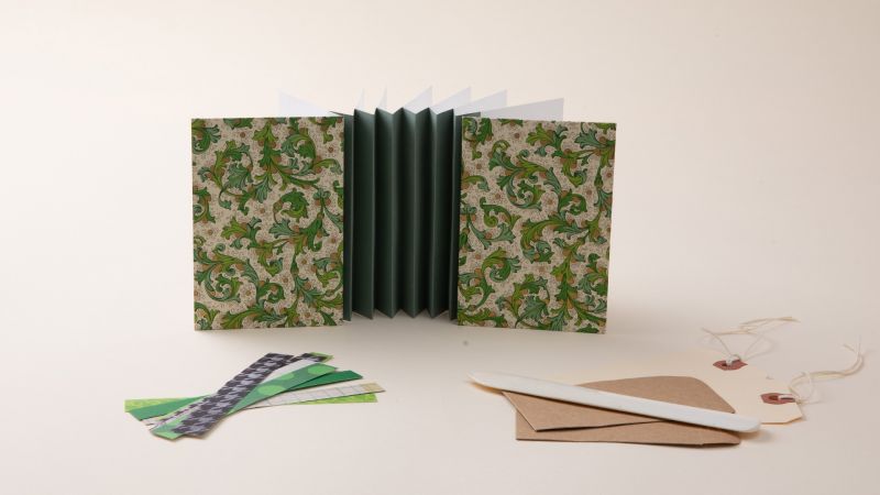 Accordion-style notebook with a green floral cover, paper strips, envelopes, a bone folder, and string on a light background.