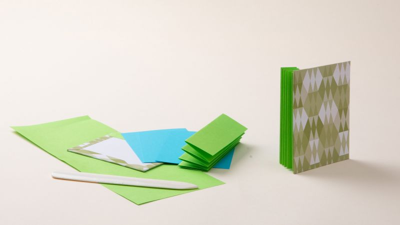 Origami papers and a bone folder on a table; a completed folded book in green and white patterns is to the right.