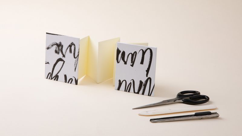 Accordion-style paper cards next to scissors, a craft knife, and a bone folder on a light surface.
