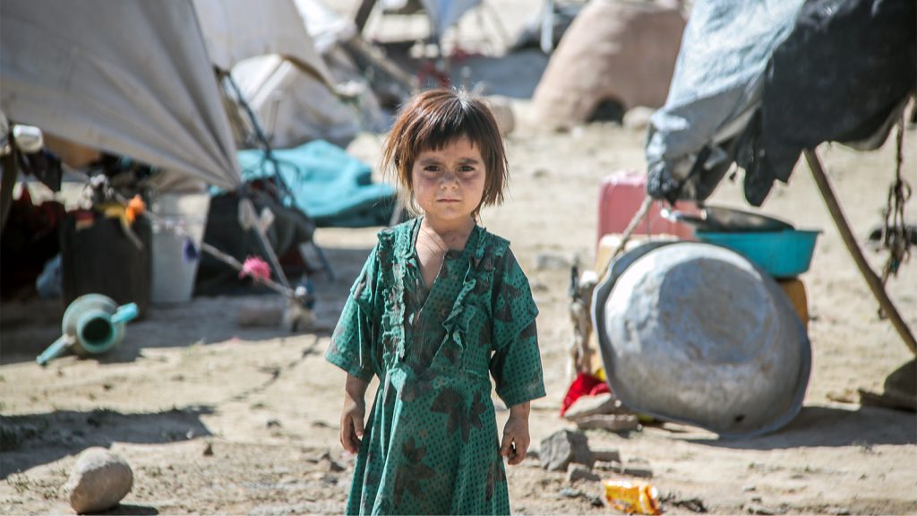 A young child in a green dress stands in an outdoor area with tents and scattered objects in the background.