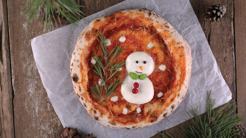 Pizza topped with cheese shaped like a snowman, herbs, and red accents, on parchment paper and wood.