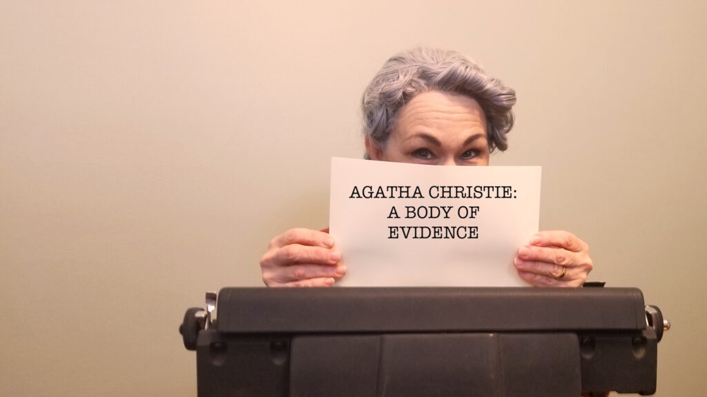 Woman with gray hair holds a "Agatha Christie: A Body Of Evidence" sign behind a vintage typewriter.