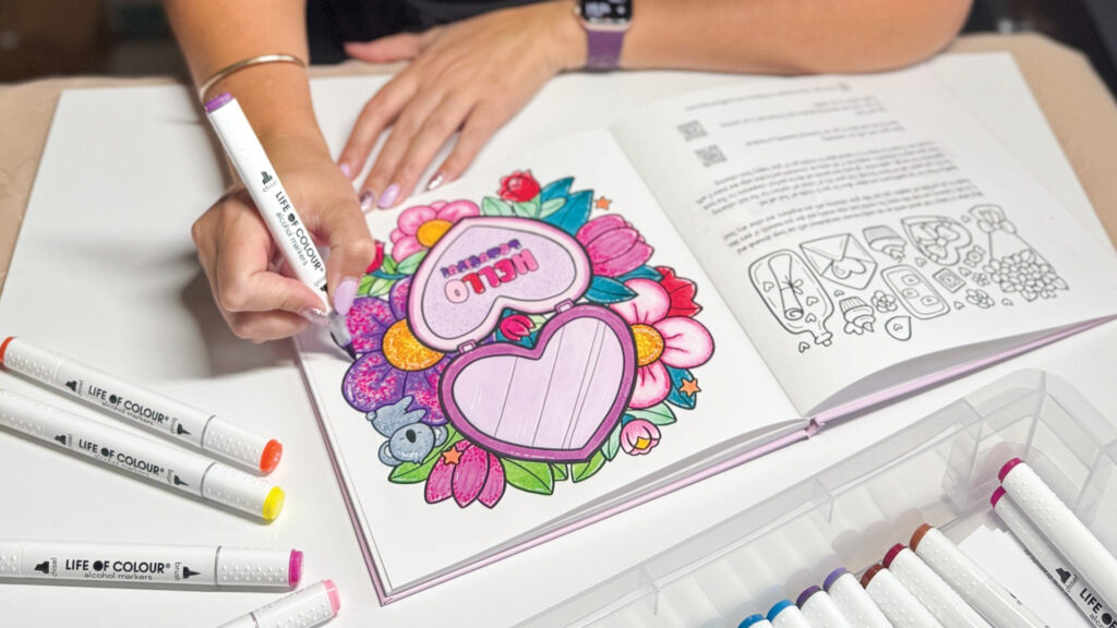 A person colors a floral heart design in an adult coloring book with bright markers on a table.