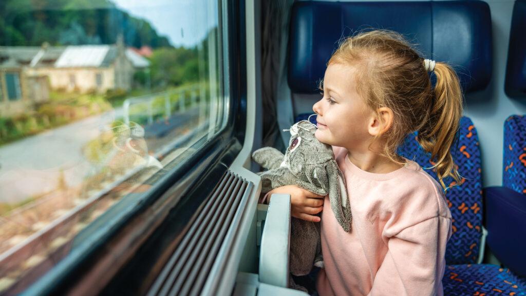 Young girl holding a stuffed animal, smiling and looking out the window while riding on a train.