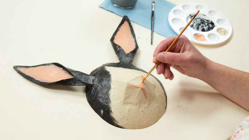 A person painting a donkey mask on a table with a palette and paintbrush beside it.