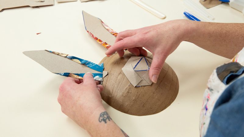 Person crafting a paper mache animal head with cardboard ears on a table.