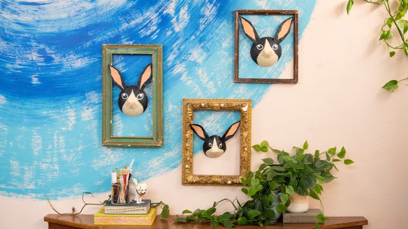 Three framed rabbit face decorations on a blue-painted wall, with a plant and pencils on a wooden table below.