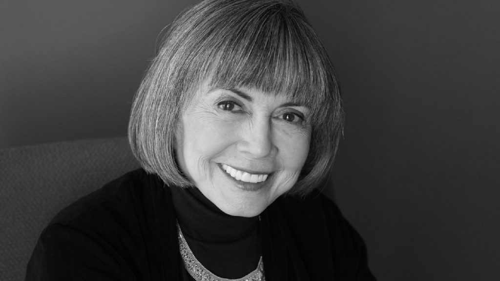 Smiling person with short, straight hair and bangs, wearing a dark outfit and necklace, posing against a plain background.